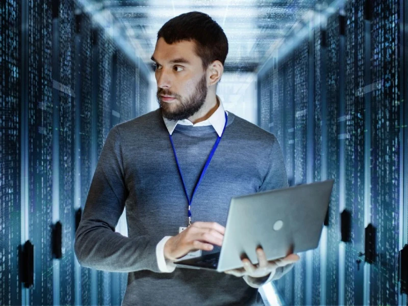         A bearded man in a gray sweater and blue lanyard is holding a laptop, standing in a server room filled with rows of data servers, ensuring the company's systems remain secure and operational while minding the importance of Worker's Compensation Insurance for all employees.